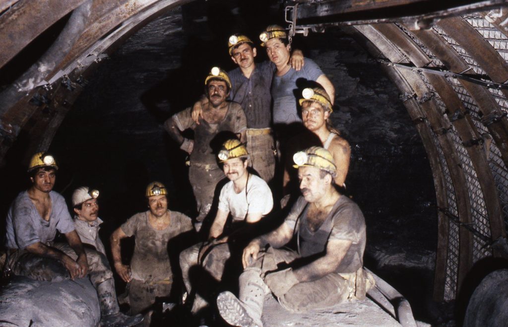 Photograph showing miners underground in dark tunnel wearing helmets and torch lights.