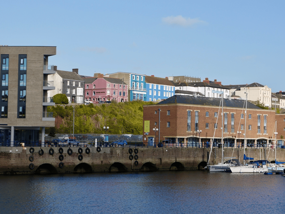 A view of the waterfront at Milford Haven on a sunny day, with boats moored in the foreground and commercial and residential properties in the background.